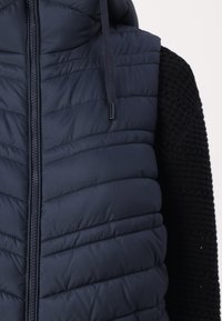 Marine blauwe gewatteerde vest met ritssluiting en capuchon. Bevat horizontale stiksels, contrasterende zwarte gebreide mouwen en trekkoordaccenten.