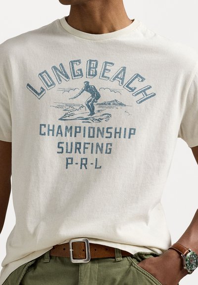 Šviesiai kreminė marškinė su mėlynu grafiniu atvaizdu, vaizduojančiu banglentininką, užrašais „LONG BEACH CHAMPIONSHIP SURFING P-R-L“ ir susiraukšlėtu apykaklės dizainu.