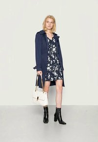 Casaco trench azul sobre um vestido floral azul. Botas de salto pretas e uma mala acolchoada branca com detalhes em dourado. Texturas suaves e formas definidas.