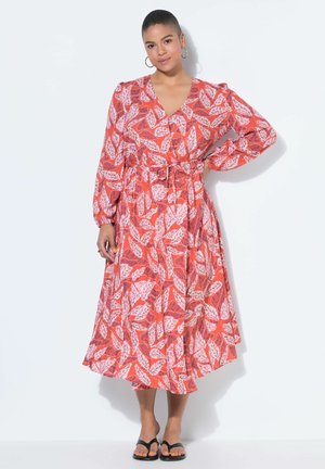 Femme aux cheveux courts portant une robe mi-longue à manches longues à motif de feuilles rouges et roses, debout avec une main sur la hanche et des sandales noires.