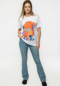 White cotton t-shirt featuring a colorful graphic print of a face with orange and blue accents. Paired with light blue flared jeans and black shoes.