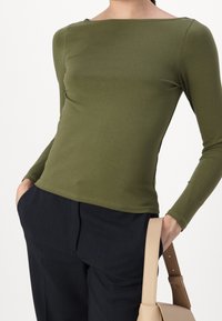 Långärmad topp i olivgrön, med båturringning och figurnära design. Matches med marinblå byxor och en beige handväska. Slät textur.