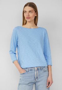 Hellblaues, langärmliges Oberteil aus einem weichen, strukturierten Stoff mit einem weiten Ausschnitt, kombiniert mit hellen Jeans aus Denim mit einem Kordelzugbund.