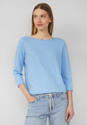 Light blue, long-sleeve top in a soft, textured fabric with a wide neckline, paired with light denim jeans featuring a drawstring waist.