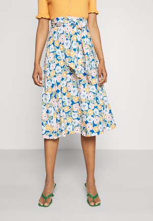 Femme portant une jupe fleurie mi-longue avec des fleurs bleues, roses et jaunes, un haut jaune côtelé et des sandales vertes ouvertes aux orteils.
