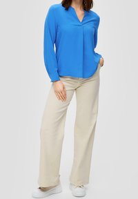 Blouse bleue à manches longues avec un décolleté en V, associée à un pantalon large beige clair et des baskets blanches. Tissu lisse, coupe décontractée.