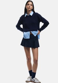 Merino villasta valmistettu tummansininen lyhyt neulepaita, vaaleansininen kauluspaita sen alla, pleated-tyylinen tummansininen hame ja valkoiset tennarit tummilla yksityiskohdilla.