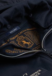 Interno di una giacca navy con una grafica gialla che presenta un emblema di un cavallo coronato e testo. Dettaglio della cerniera sul bordo e texture del tessuto visibile.