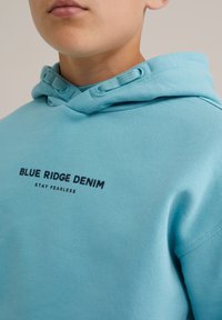 Primo piano di una persona che indossa un hoodie blu chiaro con la scritta "BLUE RIDGE DENIM STAY FEARLESS" stampata sul petto.