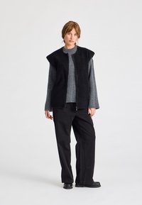 Kvinna står i svarta vida byxor, svart zip-up väst över grå stickad tröja, och svarta skor mot en helt vit bakgrund.