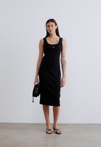 Μαύρο αμάνικο midi φόρεμα με εφαρμοστή γραμμή, υφασμάτινη υφή, με μικρό λογότυπο στο στήθος και συνδυάζεται με μαύρη τσάντα pouch και σανδάλια.