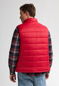 Rote, gepolsterte Weste mit hohem Kragen und gestepptem Design, getragen über einem karierten Flanellhemd. Der Stoff wirkt leicht und glatt.