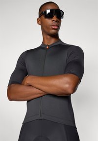 Maglia da ciclismo nera a maniche corte con finitura testurizzata, zip frontale, linguetta arancione e vestibilità aderente, abbinata a occhiali da sole neri oversize.