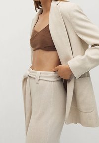Blazer beige ajusté sur un crop top marron, associé à un pantalon taille haute beige avec une ceinture en tissu et des poches latérales.