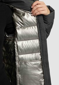 Must pufferjope läikiva hõbedase sisevoodriga; sellel on ruutõmblustega sektsioonid ja siseservas asuv tasku, mis paistab ava lähedal.