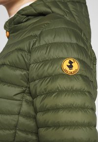 Doudoune vert olive avec texture matelassée, dotée d'une capuche et d'un écusson rond jaune sur la manche gauche.