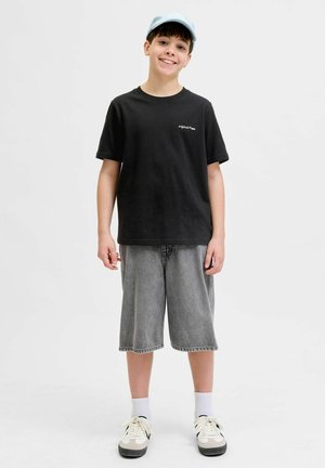 Smilende dreng iført en lyseblå kasket, sort t-shirt, grå knælange shorts, hvide sokker og beige sneakers mod en ensfarvet hvid baggrund.