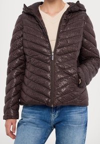 Veste chauffante matelassée marron avec une texture brillante, motif de couture horizontale, fermeture éclair et capuche. Portée sur un léger pull en V.