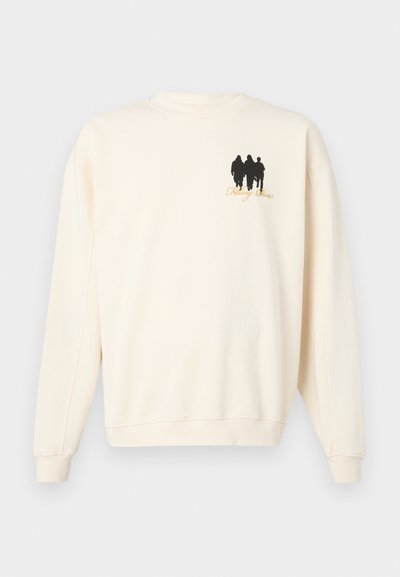 Kremna dukserica iz mehkega materiala. Vsebuje črno grafiko treh figur na levem prsnem predelu in napis "Fitting Dress" v zlati barvi.