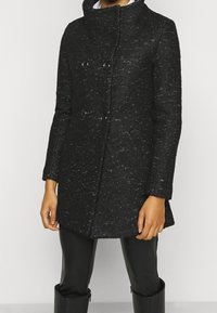 Svart dubbelknäppt kappa med en texturerad väv, som har hög krage, stora svarta knappar och sidofickor. Matchad med svarta byxor.