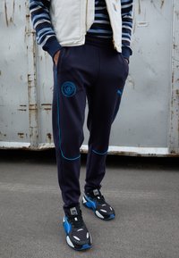 Jūras spēku sporta bikses ar gaiši ziliem akcentiem un Mančestras City logo, apvienotas ar melniem un ziliem Puma sporta apaviem, kas izceļas ar sieta dizainu.