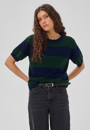 Giovane donna con lunghi capelli ricci che indossa un maglione a maniche corte a righe verde e blu navy e jeans neri con una cintura nera.