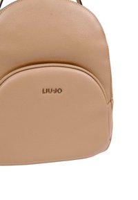 Beige ryggsäck i läder med rundad form, dragkedjestängning och en framficka. Har guldfärgad märkning med "LIU JO" på framsidan.