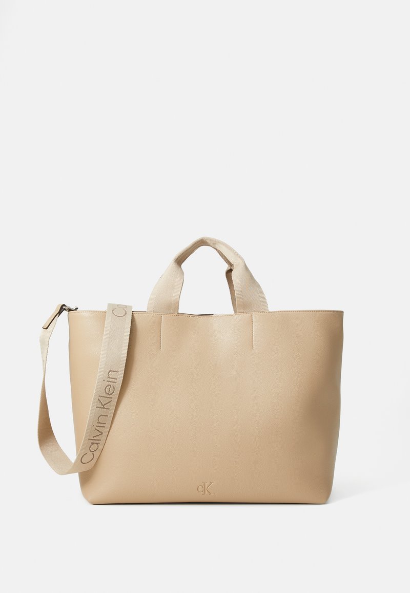 Beige lærveske med doble håndtak på toppen og en avtakbar stropp. Har minimalistisk design og preget logo. Glatt tekstur, uten dekorasjoner.