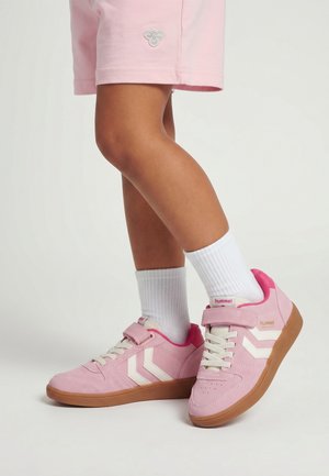 Kind draagt roze sneakers met witte chevronstrepen, witte sokken en roze korte broek, staand op een witte ondergrond.