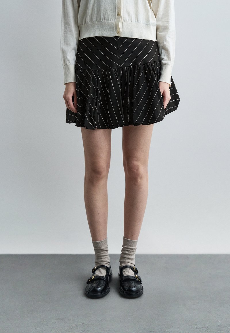 Άτομο με μπεζ ζακέτα, μαύρη μίνι φούστα με διαγώνιες ρίγες, μπεζ κάλτσες και μαύρα παπούτσια με αγκράφα που στέκεται σε γκρι δάπεδο.