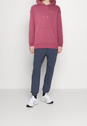 Person trägt einen mauvefarbenen Hoodie, marineblaue Jogginghosen, schwarze Socken und weiße Laufschuhe, steht vor einem einfarbigen hellen Hintergrund.