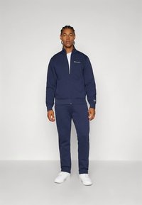 Granatowy dres wykonany z teksturowanej tkaniny; kurtka na zamek z mankietami na rękawach, pasujące spodnie oraz białe trampki o minimalistycznym wzornictwie.