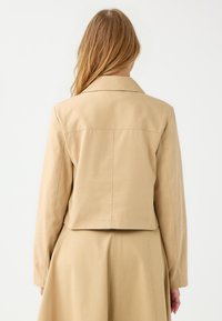 Beige Cropped-Jacke mit klassischem Kragen, langen Ärmeln und glattem Stoff. Fällt durch Rückennaht und ein schlichtes, unverziertes Design auf.