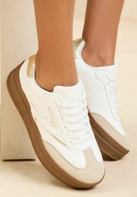 Adidași albi cu un superior din piele netedă, accente din suede bej, detaliu auriu pe toc, vârf rotund și talpă din cauciuc maro.