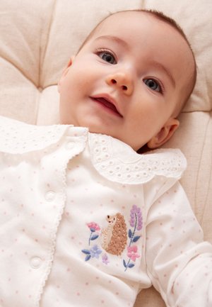 Usmievavé bábätko ležiace v bielom oblečení s vyšitým ježkom a farebnými kvetmi na hrudi.