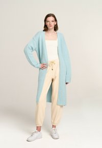Light blue knitted cardigan with long sleeves and open front, worn over a white top and cream tapered pants. White sneakers complete the outfit.