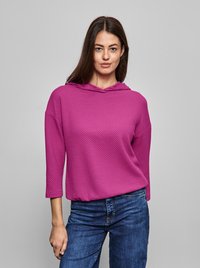 Sudadera rosa con capucha y un patrón texturizado, mangas tres cuartos y un corte relajado, combinada con jeans azules. Fondo neutro.
