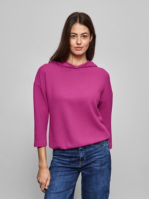 Sweatshirt à capuche rose avec un motif texturé, manches trois quarts et coupe décontractée, associé à un jean bleu. Fond neutre.