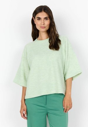 Woman with long dark hair wearing a light green oversized sweater and matching green pants, standing against a white background.