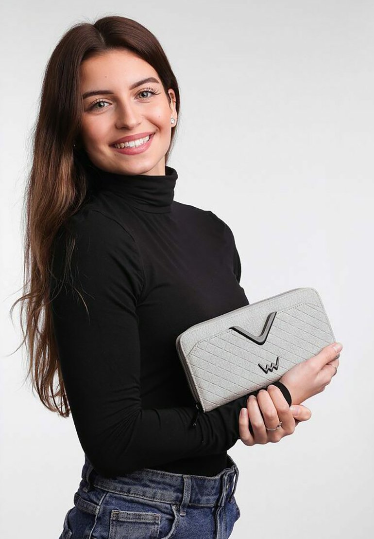 Šedá prošívaná peněženka s elegantním designem, která disponuje černým akcentem a zipovým uzávěrem, držena v ruce na neutrálním pozadí.