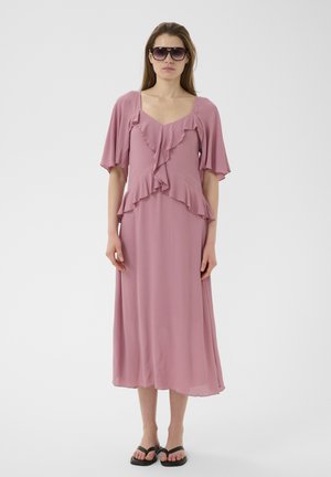 Femeie îmbrăcată într-o rochie midi malva cu volane, mâneci evazate, papuci negri și ochelari de soare supradimensionați negri, stând în fața unui fundal simplu.