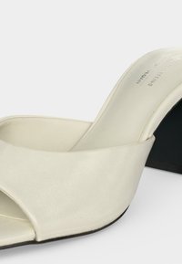 Λευκό mule με ανοιχτά δάχτυλα, χαμηλό χοντρό τακούνι και λείο δερμάτινο επάνω μέρος, εμφανίζεται από μπροστινή πλευρική γωνία.