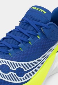Sapatilha atlética azul com parte superior em malha, superfície texturizada, detalhes em branco e amarelo, sistema de atacadores proeminente e padrão distintivo na lateral.