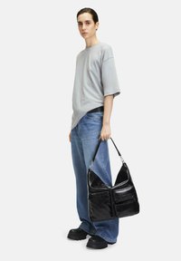 Czarna skórzana torba hobo o błyszczącej teksturze, z dwiema przednimi kieszeniami na zamek i zaokrąglonym topem, trzymana przez osobę ubraną w szary, oversize'owy t-shirt i niebieskie dżinsy.