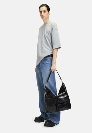 Schwarze Leder-Hobo-Tasche mit glänzender Textur, zwei vorderen Reißverschlusstaschen und einer geschwungenen Oberseite, gehalten von einer Person, die ein graues Oversized-T-Shirt und blaue Jeans trägt.