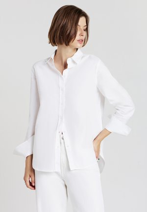 Woman with short brown hair wearing a white button-up shirt and white pants, posing with one hand in pocket, looking to her left.