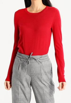 Femme portant un pull rouge à manches longues et un pantalon gris à carreaux taille haute avec cordon à la taille.