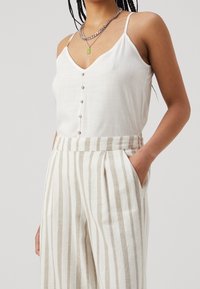 Camisole en lin blanc avec de petits boutons à l'avant, associé à un pantalon taille haute rayé en beige et blanc. Il présente des bretelles fines et des poches.