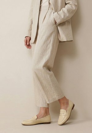 Person iført beige linnedbukser, matchende blazer og cremefarvede loafers, der står mod en lys beige væg.