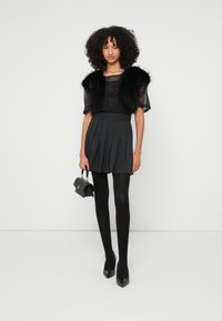 Colete de imitação de pele preta por cima de uma blusa preta transparente, combinado com uma saia cinzenta plissada, collants pretos e saltos com biqueira pontiaguda, segurando uma pequena mala.